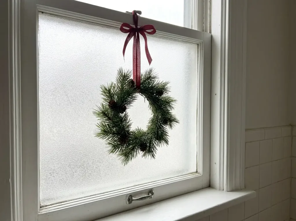 Small evergreen Christmas wreath with red ribbon hanging inside a frosted bathroom window.