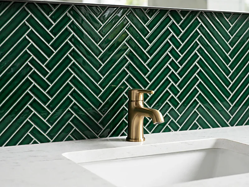 Green herringbone ceramic tile backsplash behind a white sink with brass faucet and walnut vanity.